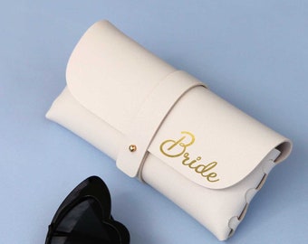 May include: White sunglasses case with the word "Bride" written in gold lettering. The case is closed and lying on a light blue surface. Next to the case are black heart-shaped sunglasses.