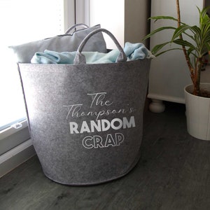 May include: A gray felt storage bin with the text "The Thompson's RANDOM CRAP" printed on the front. The bin is filled with blankets and pillows.