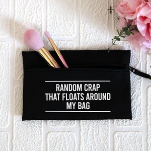 May include: Black zippered pouch with the text "RANDOM CRAP THAT FLOATS AROUND MY BAG" in white. Two makeup brushes are sticking out of the top of the pouch.