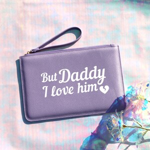 May include: A lavender-colored clutch purse with a zipper closure and a wrist strap. The purse has white text that reads "But Daddy I love him" with a small white heart.