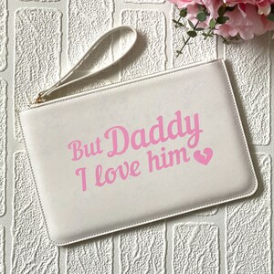 May include: White clutch bag with a pink inscription that reads "But Daddy I love him" with a pink heart.