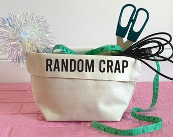 May include: A white canvas storage bin with the text "RANDOM CRAP" printed on the front. The bin is filled with various items, including green measuring tape, a pair of teal scissors, and a black cord.