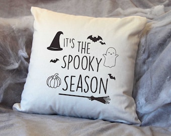 May include: White cushion with black text that reads "It's the spooky season" with a witch's hat, a ghost, a pumpkin, bats, and a broom.