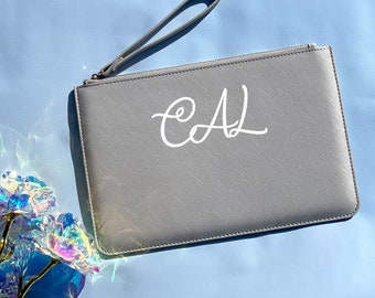 May include: A gray clutch purse with a zipper closure and a wrist strap. The purse has the initials "CAL" written in white cursive on the front.