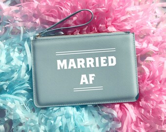 May include: A light blue clutch bag with a zipper closure and a white strap. The bag has the text "MARRIED AF" printed on the front in white.