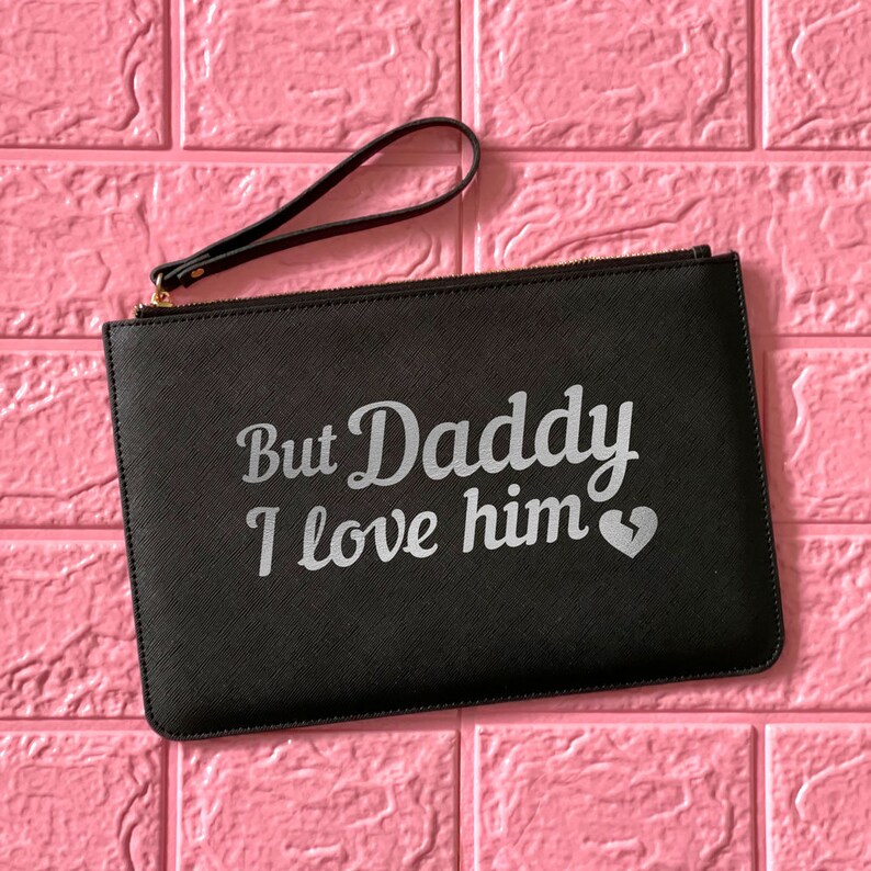 May include: Black clutch bag with a zipper closure and a silver handle. The bag has the text "But Daddy I love him" with a broken heart symbol.