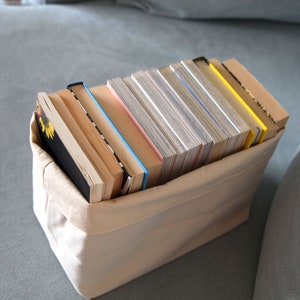 May include: A beige canvas storage bin filled with a stack of various sized books. The books are mostly white and cream with some having blue, pink, and yellow accents.