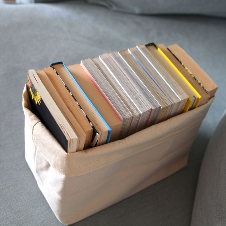 May include: A beige canvas storage bin filled with a stack of various sized books. The books are mostly white and cream with some having blue, pink, and yellow accents.