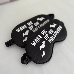 May include: Two black sleep masks with white elastic bands. The masks have white bats and the text "Wake me up on Halloween" printed on them.