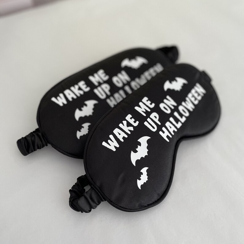 May include: Two black sleep masks with white elastic bands. The masks have white bats and the text "Wake me up on Halloween" printed on them.