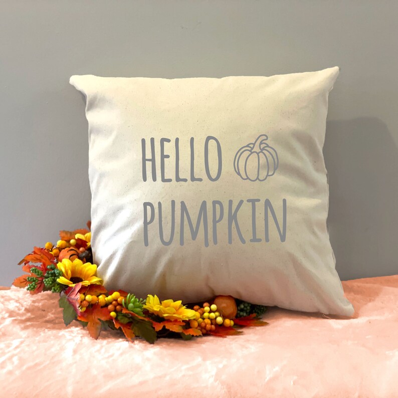 May include: A white throw pillow with the text "HELLO PUMPKIN" in gray lettering and a gray pumpkin graphic. The pillow is sitting on a pink surface with a fall-themed wreath.