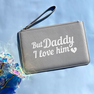 May include: A gray clutch purse with a zipper closure and a white handle. The purse has the text "But Daddy I love him" printed on the front in white.