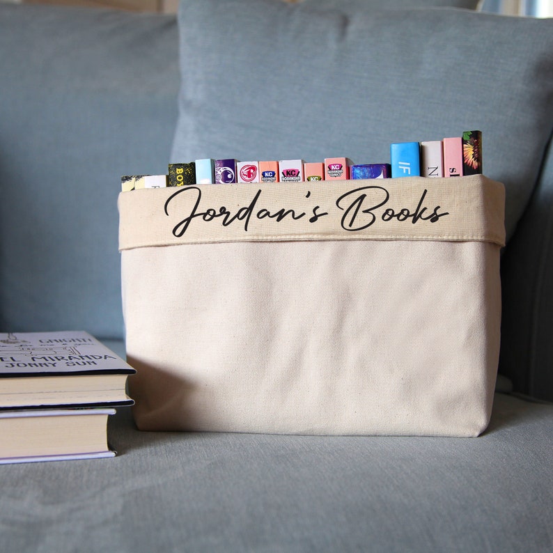 May include: A white canvas book storage bin with the text "Jordan's Books" printed on the front. The bin is filled with a variety of colorful books.
