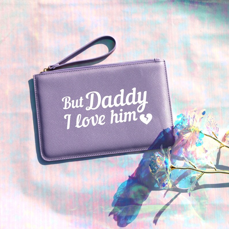 May include: A lavender-colored clutch purse with a zipper closure and a wrist strap. The purse has white text that reads "But Daddy I love him" with a small white heart.