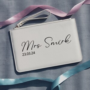 May include: White leather clutch bag with a zipper closure and a wrist strap. The bag has the text "Mrs Smirk" and the date "23.03.24" printed in black.