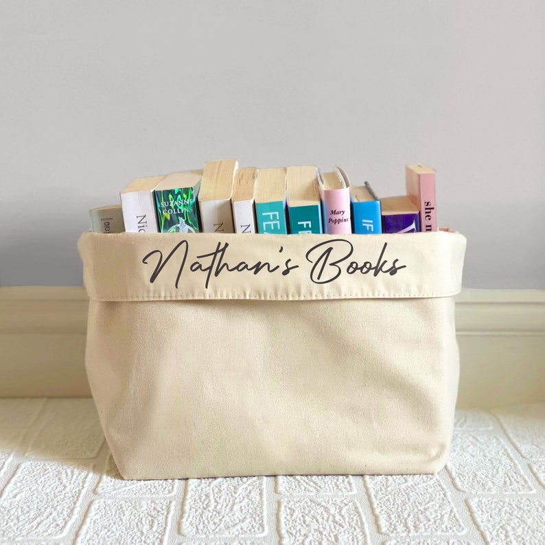 Personalised book basket. Cream colour with black script text featuring the name followed by books. The basket is full of 11 paperback books.