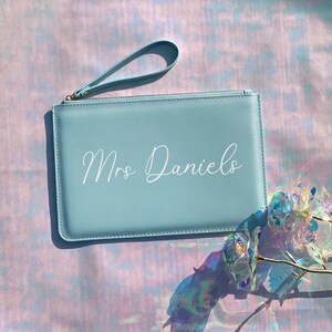 May include: A light blue clutch purse with a zipper closure and a wrist strap. The purse has the text "Mrs. Daniels" printed in white on the front.