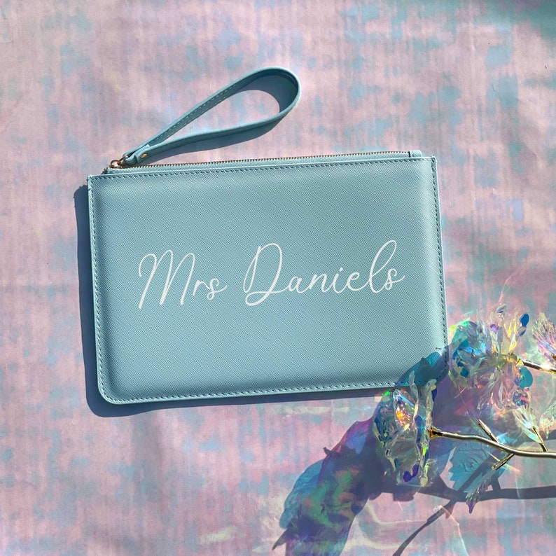 May include: A light blue clutch purse with a zipper closure and a wrist strap. The purse has the text "Mrs. Daniels" printed in white on the front.