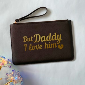 May include: A brown leather clutch with a gold zipper and a gold inscription that reads "But Daddy I love him" with a gold broken heart.