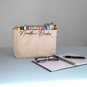 May include: A beige canvas storage bin with the text "Noelle's Books" printed on the front. The bin is filled with a variety of books. A notebook with a black cover and a black pen are on a light blue surface in front of the bin.