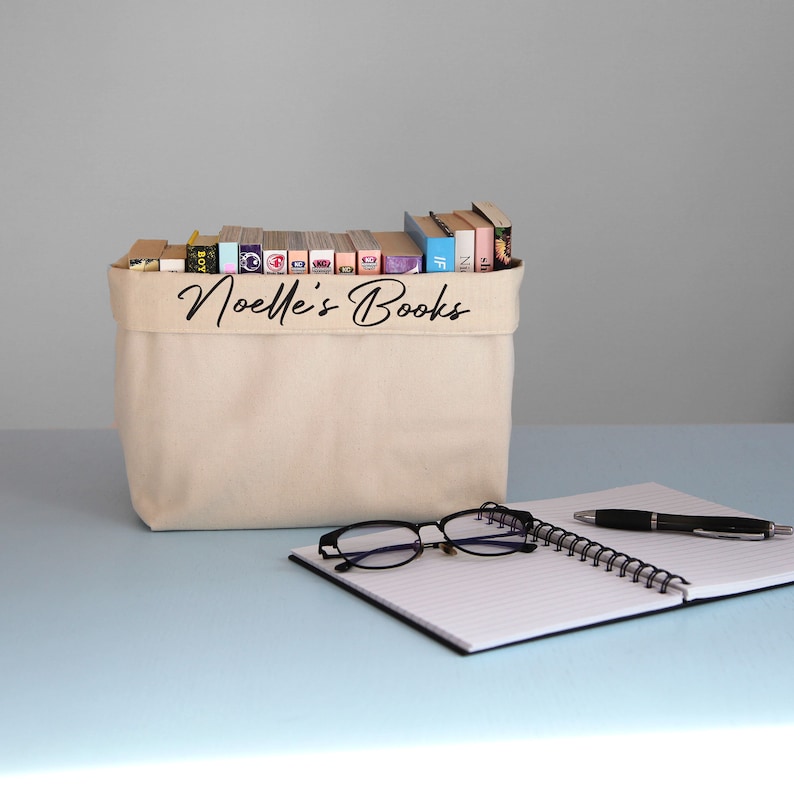 May include: A beige canvas storage bin with the text "Noelle's Books" printed on the front. The bin is filled with a variety of books. A notebook with a black cover and a black pen are on a light blue surface in front of the bin.