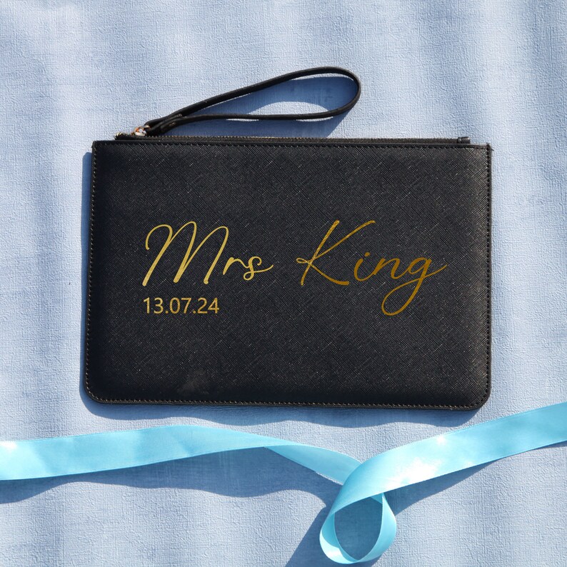 May include: Black clutch bag with a gold zipper and a gold inscription that reads "Mrs King 13.07.24".