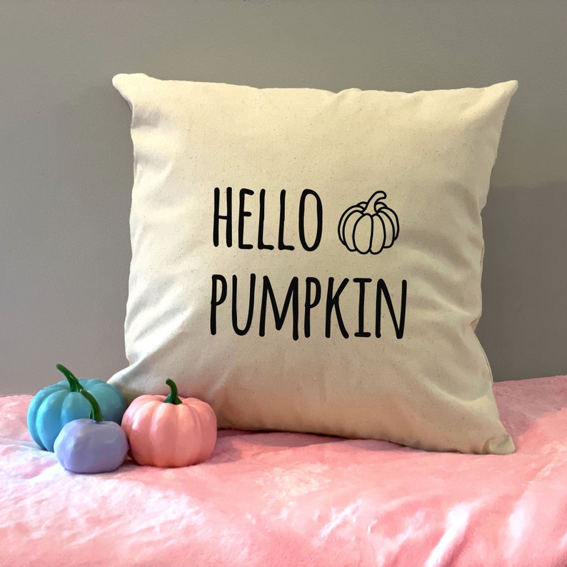 May include: A white throw pillow with black text that reads "HELLO PUMPKIN" and a black outline of a pumpkin. The pillow is sitting on a pink surface with three small decorative pumpkins.