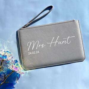 May include: A gray clutch purse with a zipper closure and a wrist strap. The purse has the text "Mrs Hunt" and "24.02.24" printed in white.