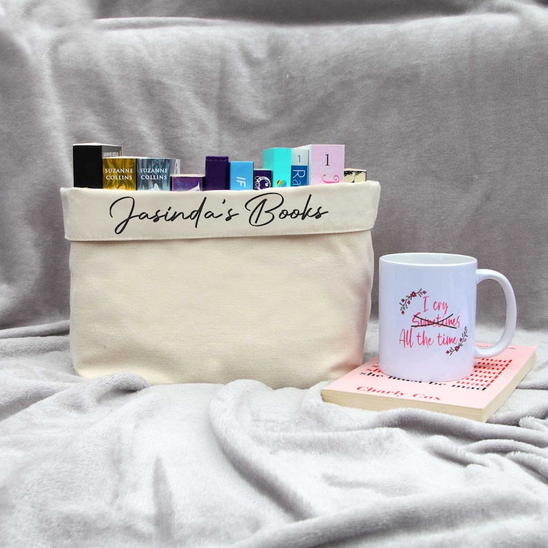 Personalised book basket. Cream colour with black script text featuring the name followed by books. The basket is full of 11 paperback books. Next to the basket is a book with a mug placed ontop.