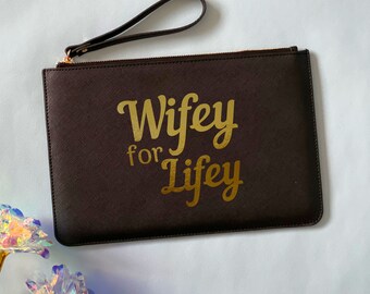 May include: A brown clutch purse with a gold zipper and a gold handle. The purse has the text "Wifey for Life" printed on the front in gold.