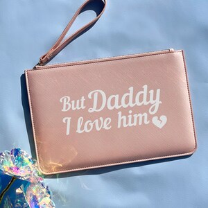 May include: A pink clutch with a zipper closure and a white strap. The clutch has the text "But Daddy I love him" printed on the front in white.