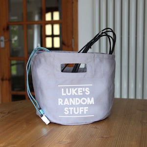 May include: A pale grey fabric storage bag with the text "LUKE'S RANDOM STUFF" in white. The bag has a rectangular opening on the top and a blue and white cord. Black cables are visible inside the bag.