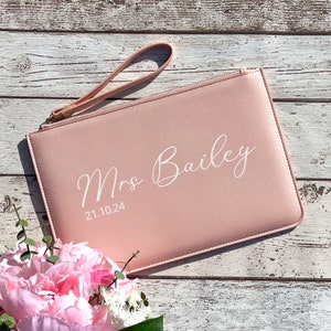 May include: A blush pink clutch with a wrist strap and the text "Mrs. Bailey 21.10.24" in white.