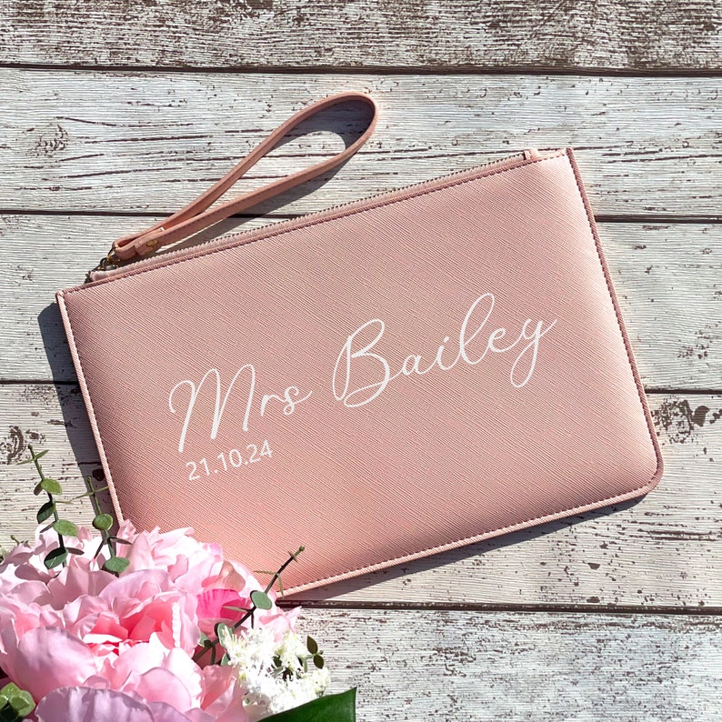 May include: A blush pink clutch with a wrist strap and the text "Mrs. Bailey 21.10.24" in white.