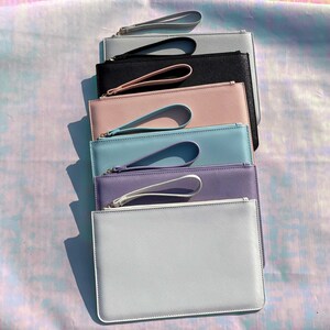 May include: A stack of seven colorful leather clutches with gold zippers and wrist straps. The clutches are arranged from lightest to darkest, with the top clutch being white, followed by light blue, lavender, light blue, pink, black, and gray.