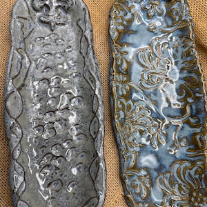 May include: Two ceramic serving trays with a distressed finish. One is gray with a fleur-de-lis design, the other is blue with a floral pattern. Both have a textured surface and are on a burlap background.
