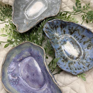 Peut inclure: Trois plats en céramique en forme d'huître dans des tons de bleu, de gris et de violet. Les plats ont une finition texturée et émaillée et sont disposés sur une surface blanche avec du feuillage vert.