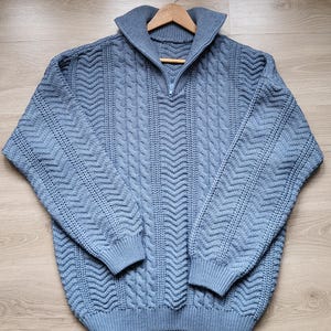 Può includere: Un maglione di lana a treccia blu chiaro con chiusura a zip a metà. Il maglione ha un collo e polsini a coste.