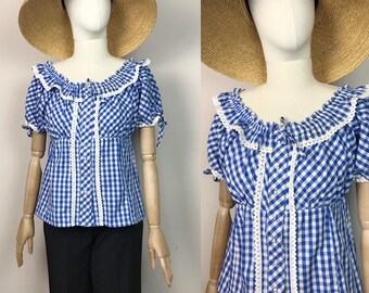 Puede incluir: Una blusa de cuadros azules y blancos con ribete de encaje blanco y cuello de volantes. La blusa tiene mangas cortas y cierre de botones en la parte delantera.