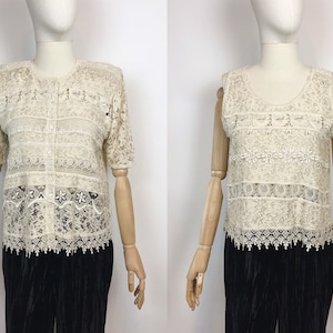 Op de afbeelding: Twee witte gehaakte tops met korte mouwen en een gegolfde zoom. De top aan de linkerkant heeft een knoopsluiting aan de voorkant, terwijl de top aan de rechterkant mouwloos is.