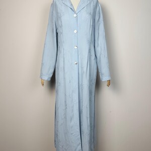 May include: A long-sleeved, floor-length light blue duster coat featuring a subtle tonal floral embroidery pattern throughout. The garment has a classic notched collar, a full-length front button closure with four visible buttons, and side slits.