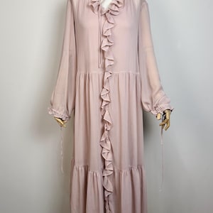 May include: A long-sleeved, dusty pink midi dress featuring a vertical ruffled trim down the center front, a round neckline with a tie closure, and tiered skirt sections. The sleeves are loose with gathered cuffs and thin hanging tie details.