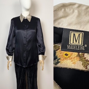 Puede incluir: Blusa de satén negro con cuello y puños en color champán en contraste. La camisa de manga larga tiene una botonadura frontal y se combina con un pantalón de terciopelo negro. La etiqueta dice "MADELEINE".
