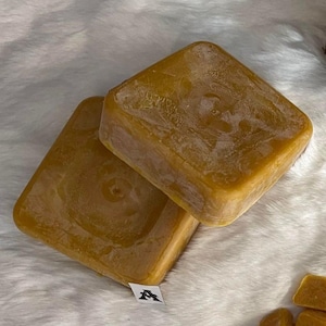 Peut inclure: Deux blocs carrés de cire d'abeille, l'un sur l'autre, tous deux de couleur jaune clair. La cire d'abeille est lisse et a une surface légèrement texturée.