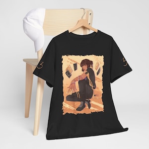 Op de afbeelding: Zwart T-shirt met een illustratie van een persoon en kaarten op een warme oranje achtergrond. Het ontwerp bevat een figuur in donkere kleding met armwikkels. Het nummer "3" is in het oranje op de mouw gedrukt.