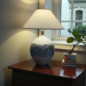 May include: A ceramic table lamp with a cream-colored lampshade and a round, patterned base. The base features a blue and brown leaf design. The lamp sits on a dark wood nightstand, next to a small potted plant and books.