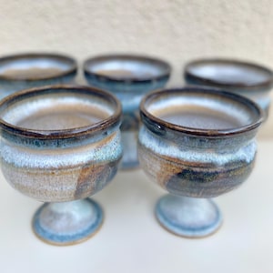 Puede incluir: Conjunto de cinco tazas de cerámica con un esmalte blanco y detalles marrones y azules. Las tazas tienen una base con pie.