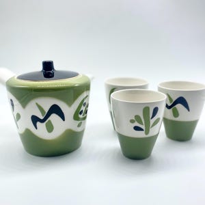 以下が含まれることがあります： 黒い蓋が付いたティーポットと、お揃いのカップ3つがセットになった陶器のティーセット。セットは主に緑と白で、ネイビーと緑の抽象的なデザインが繰り返されています。カップは緑色のベースです。