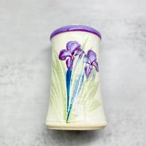 Puede incluir: Un recipiente de cerámica con borde morado y diseño floral. El recipiente presenta dos flores de iris moradas con hojas verdes y tallos azules. El fondo es blanquecino.