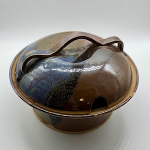 May include: A handmade ceramic lidded bowl with a brown, blue, and tan glaze. The bowl has a rounded shape with a matching lid and a curved handle. The bowl is suitable for serving or storage.